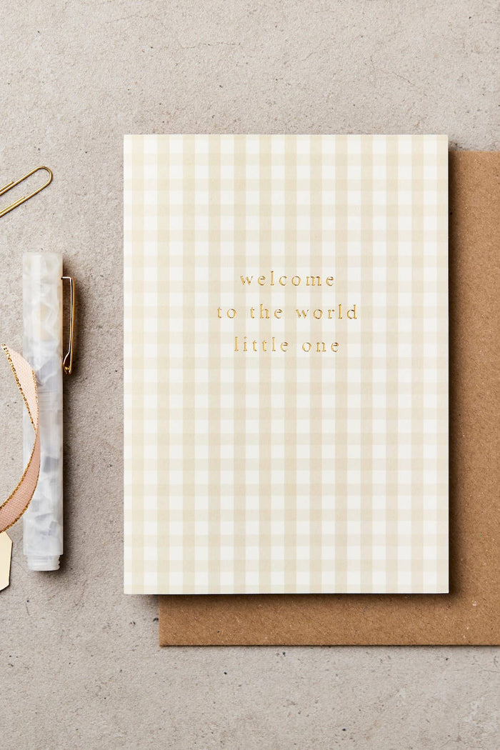 Greeting card with 'welcome to the world little one' text on a checkered pattern, with a brown envelope and stationery items on a beige background.