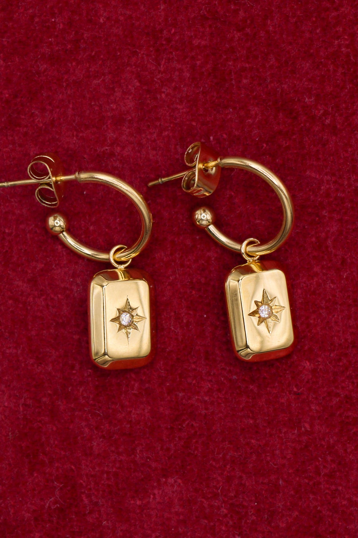A pair of gold hoop earrings with rectangular pendant charms featuring a starburst design and a small clear stone, displayed on a textured dark red velvet background.