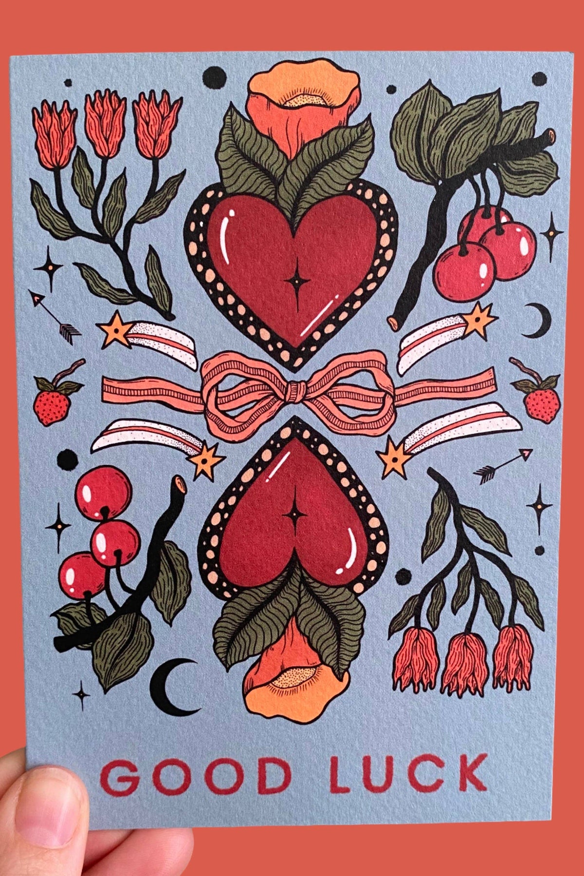Card with red hearts, flowers, and 'Good Luck' text on a blue background held against an orange background
