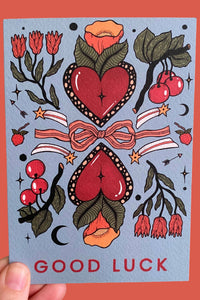 Card with red hearts, flowers, and 'Good Luck' text on a blue background held against an orange background