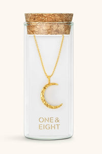 A gold crescent moon pendant necklace displayed inside a clear glass vial with a cork stopper. The vial is labeled 'ONE & EIGHT' in gold lettering on the front, set against a solid off-white background.