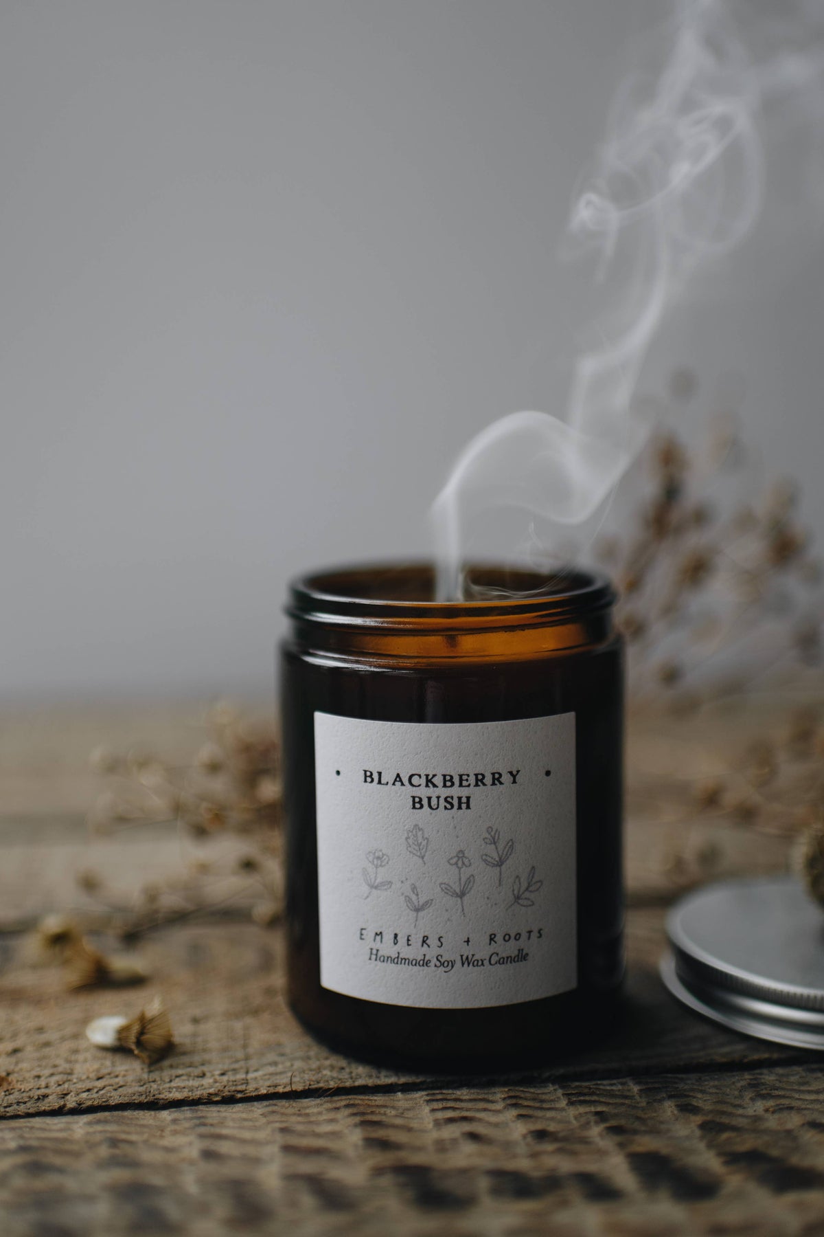 Candle labeled 'Blackberry Bush' with smoke rising, on a textured surface with dried plants.