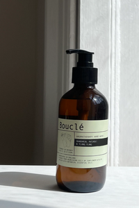 An amber glass pump bottle with a black dispenser and a cream label reading Bouclé, standing on a white surface against a white wall with strong natural light and a soft shadow.