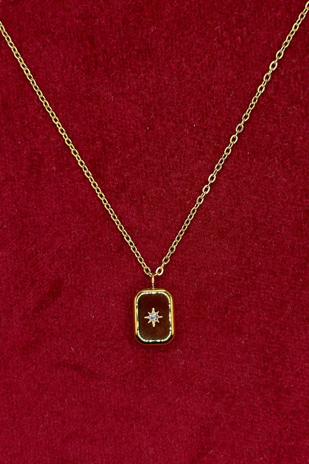 A gold chain necklace with a small rectangular pendant featuring a central star motif, displayed on a textured red velvet background.