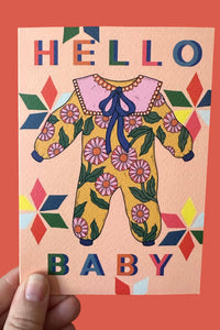 A hand holds a square greeting card featuring a retro-style illustration of a baby onesie with a pink collar and blue bow, surrounded by colorful geometric patterns and the words 'HELLO BABY'. The card is set against a solid coral-red background.