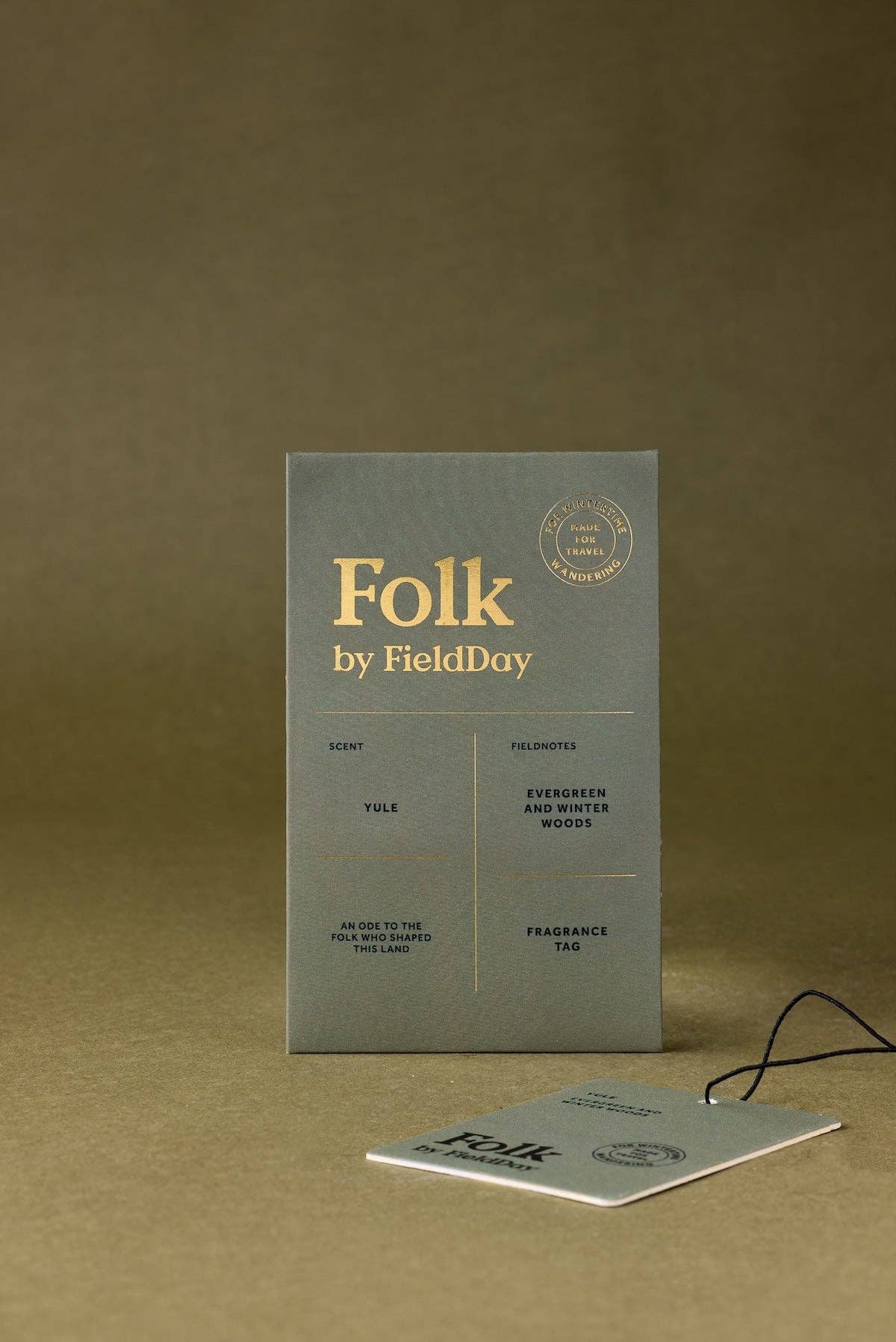 A square gray fragrance tag for 'Folk by FieldDay' stands upright on a warm brown background, with a smaller matching card placed in front and a thin black string attached. The text is printed in a muted gold tone.