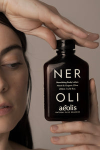 A close-up lifestyle shot of a woman holding a dark amber bottle of aloe vera body lotion. The bottle features white text reading 'NER OLI aeolis' and is held against a neutral, warm beige background.