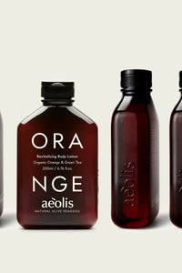 A row of amber glass body care bottles on a light warm-gray background. The center bottle is shown front-facing with large white text reading 'ORA NGE' and smaller text 'Revitalising Body Lotion' plus the brand 'aeolis'.