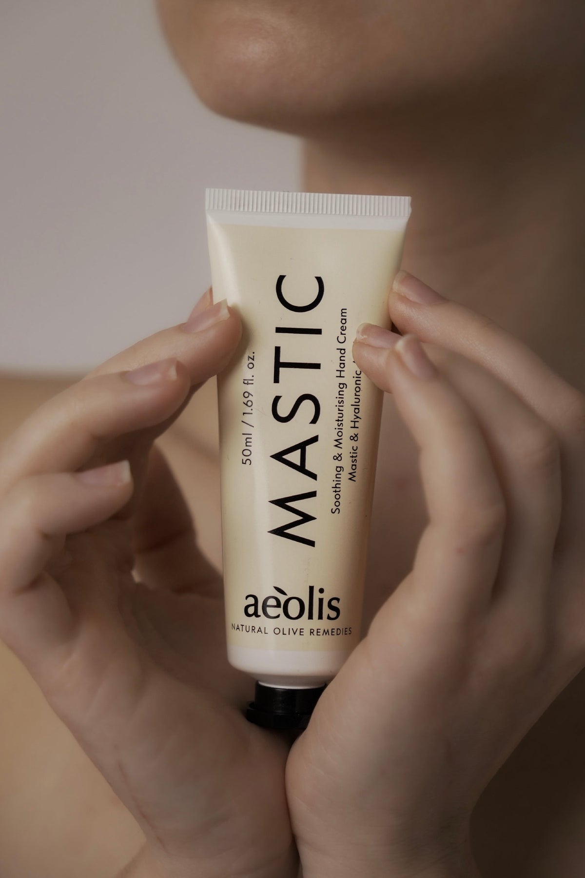 A person holds a squeeze tube of aeolis MASTIC Soothing & Moisturizing Hand Cream against their chest. The tube is a warm cream color with black text, and the background is a soft, warm beige.