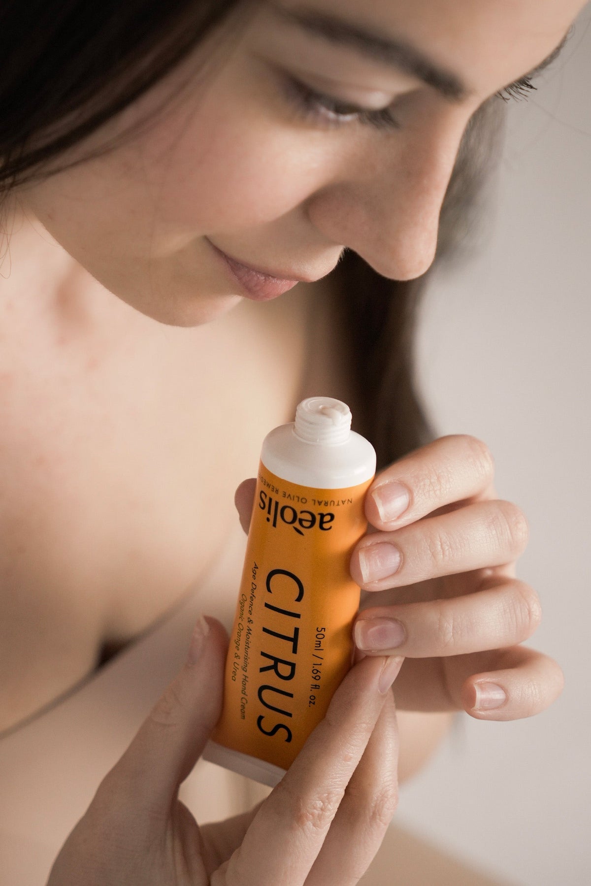 A woman holding a squeeze tube of aloe vera facial gel with a bright orange label and white cap, with her face blurred in the background.
