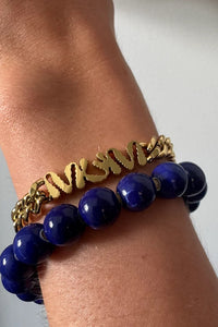 Gold chain and blue beaded bracelet on a wrist against a neutral background