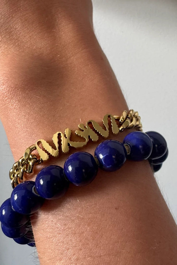 Gold chain and blue beaded bracelet on a wrist against a neutral background