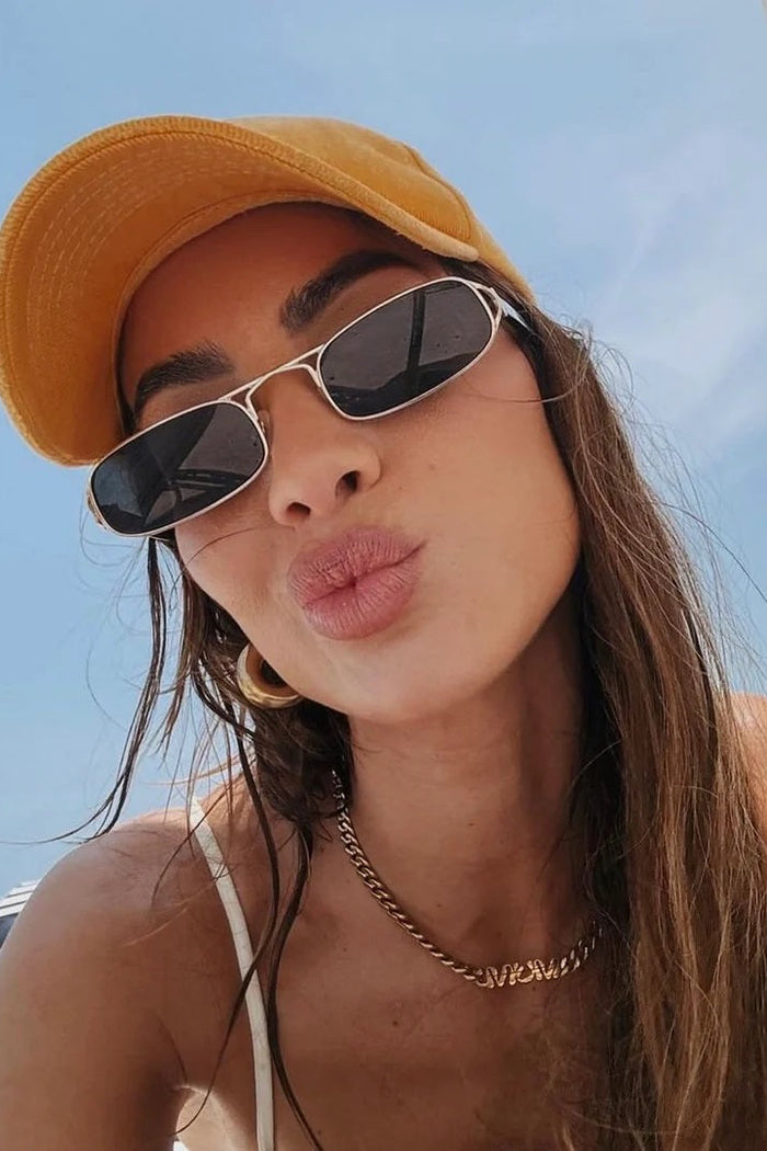 Woman wearing sunglasses and a yellow cap on a beach with a striped umbrella in the background.