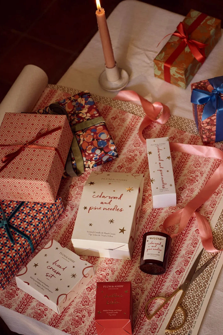 A styled flat-lay of various gift boxes, ribbons, and candles on a patterned fabric surface. Visible items include a candlestick with a lit candle, a patterned gift box with a red ribbon, a blue patterned gift box, a cream-colored box with gold stars reading 'coloured and painted', a small dark bottle, and other wrapped presents.