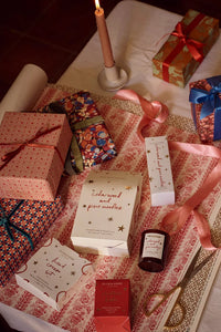 A styled flat-lay of various gift boxes, ribbons, and candles on a patterned fabric surface. Visible items include a candlestick with a lit candle, a patterned gift box with a red ribbon, a blue patterned gift box, a cream-colored box with gold stars reading 'coloured and painted', a small dark bottle, and other wrapped presents.