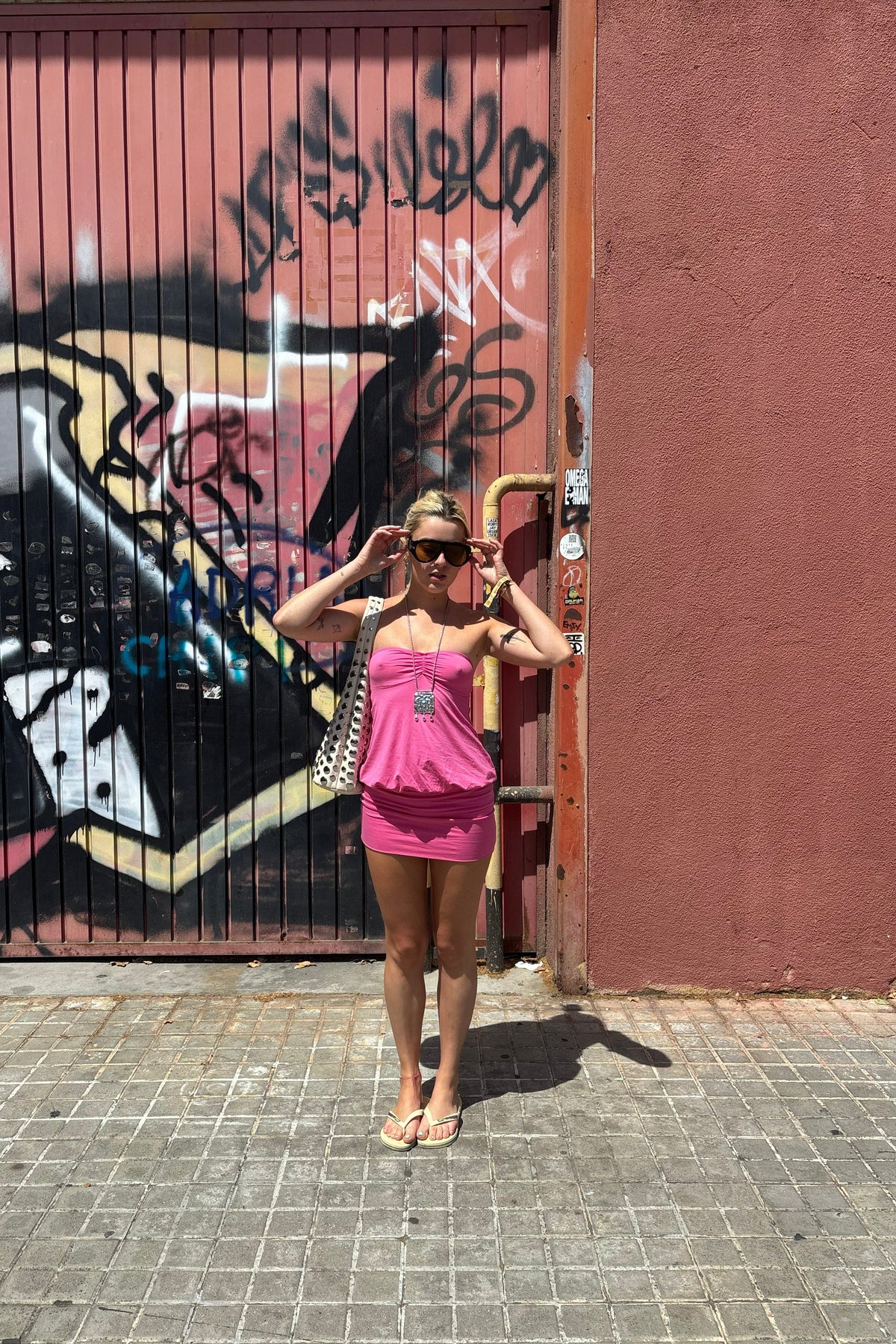 A woman wearing a pink two-piece swimsuit and sunglasses stands on a paved street in front of a graffiti-covered metal gate and a pink wall.