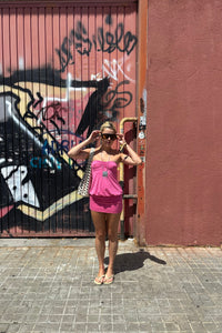 A woman wearing a pink two-piece swimsuit and sunglasses stands on a paved street in front of a graffiti-covered metal gate and a pink wall.