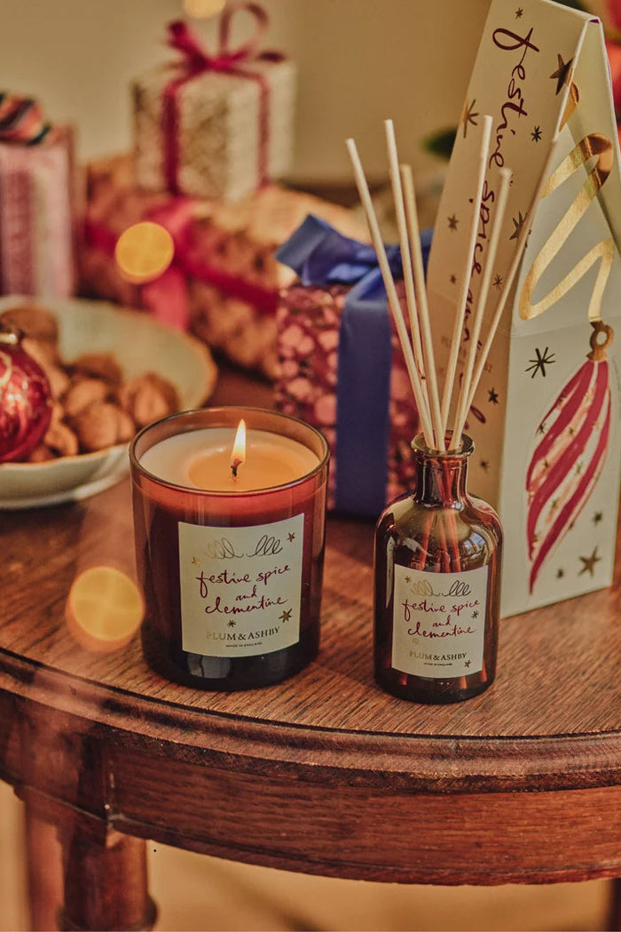 A lit scented candle in a dark amber glass jar sits on a wooden table beside a matching reed diffuser bottle with reeds and a decorative gift box. The scene is styled with wrapped presents, ornaments, and warm holiday lighting in the background.