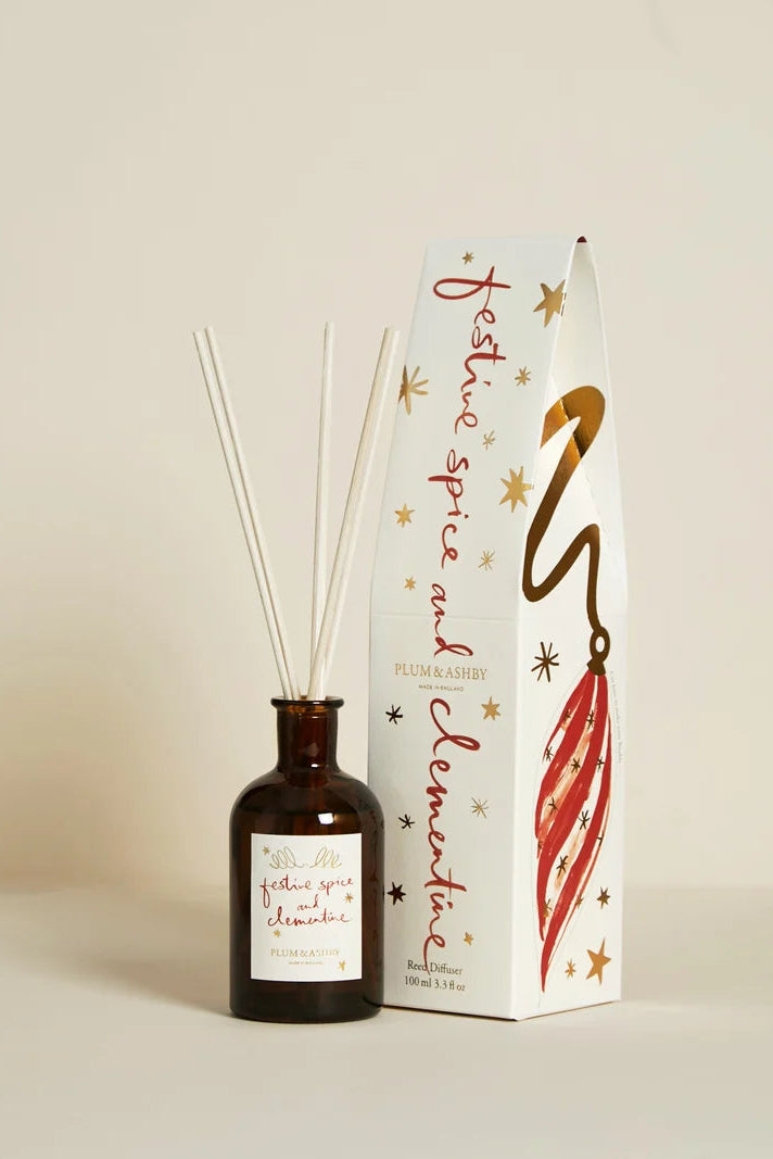 A reed diffuser bottle with white reeds next to its white packaging box decorated with gold stars and a red ornament illustration. The bottle has a white label with red text, and the box reads 'festive spice and clove' by Plum & Ash.