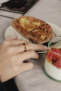 Hand with a gold ring near toast and a glass of yogurt with fruit on a table.