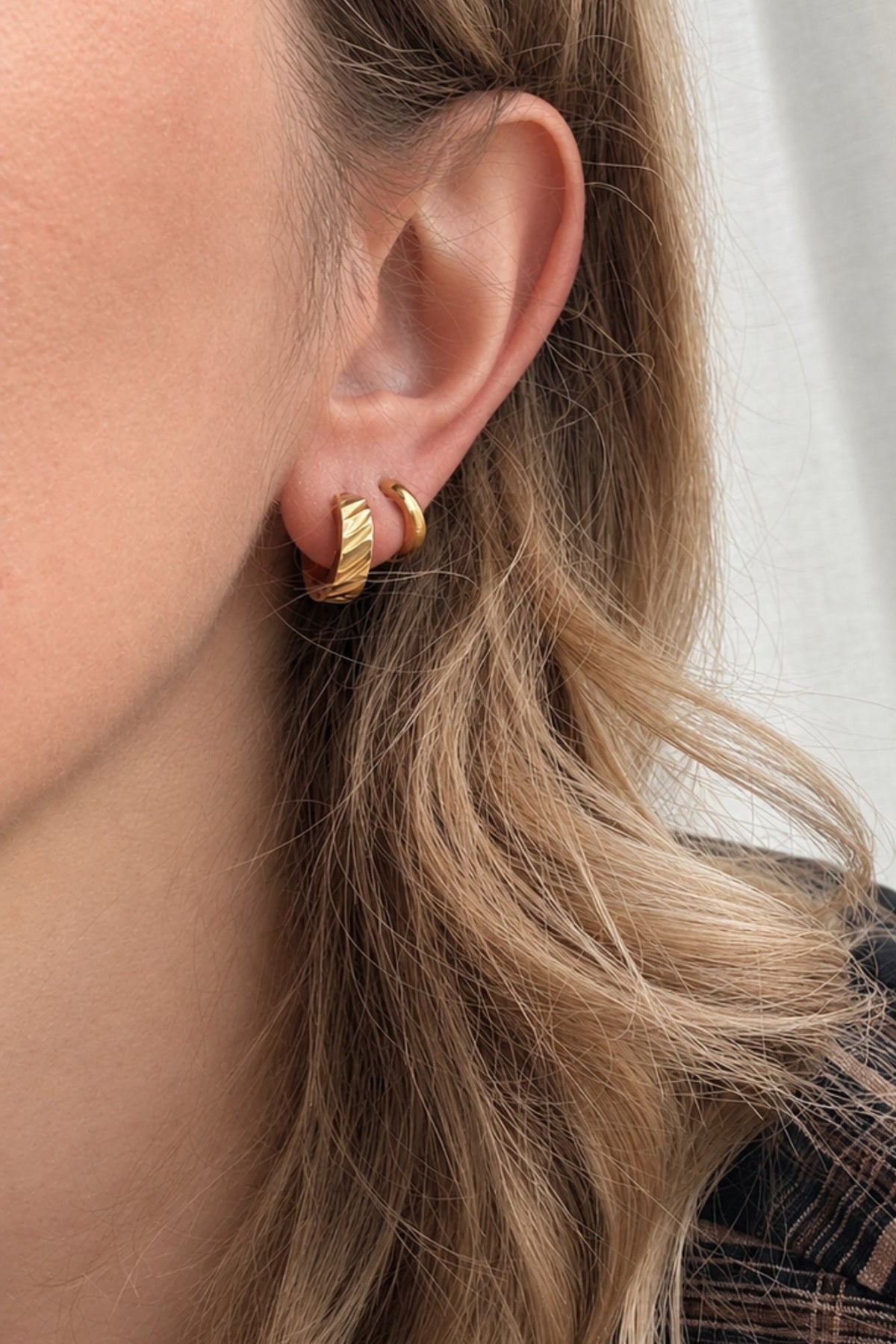 A close-up of a person's ear wearing two small gold hoop earrings, with light brown hair and a plaid shirt visible against a soft white curtain background.