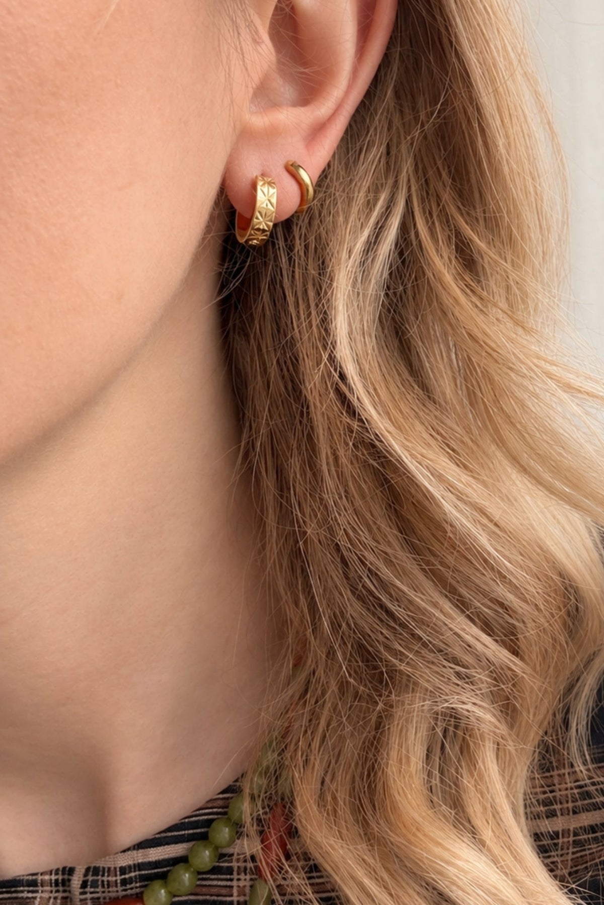 A close-up of a person's ear wearing two small gold hoop earrings, with blonde hair and a plaid top visible against a light curtain background.