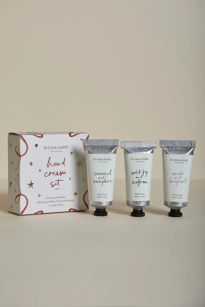 A hand cream set by Plum Ashby, featuring a decorative white and gold gift box alongside three white squeeze tubes with silver caps, arranged on a neutral beige background.