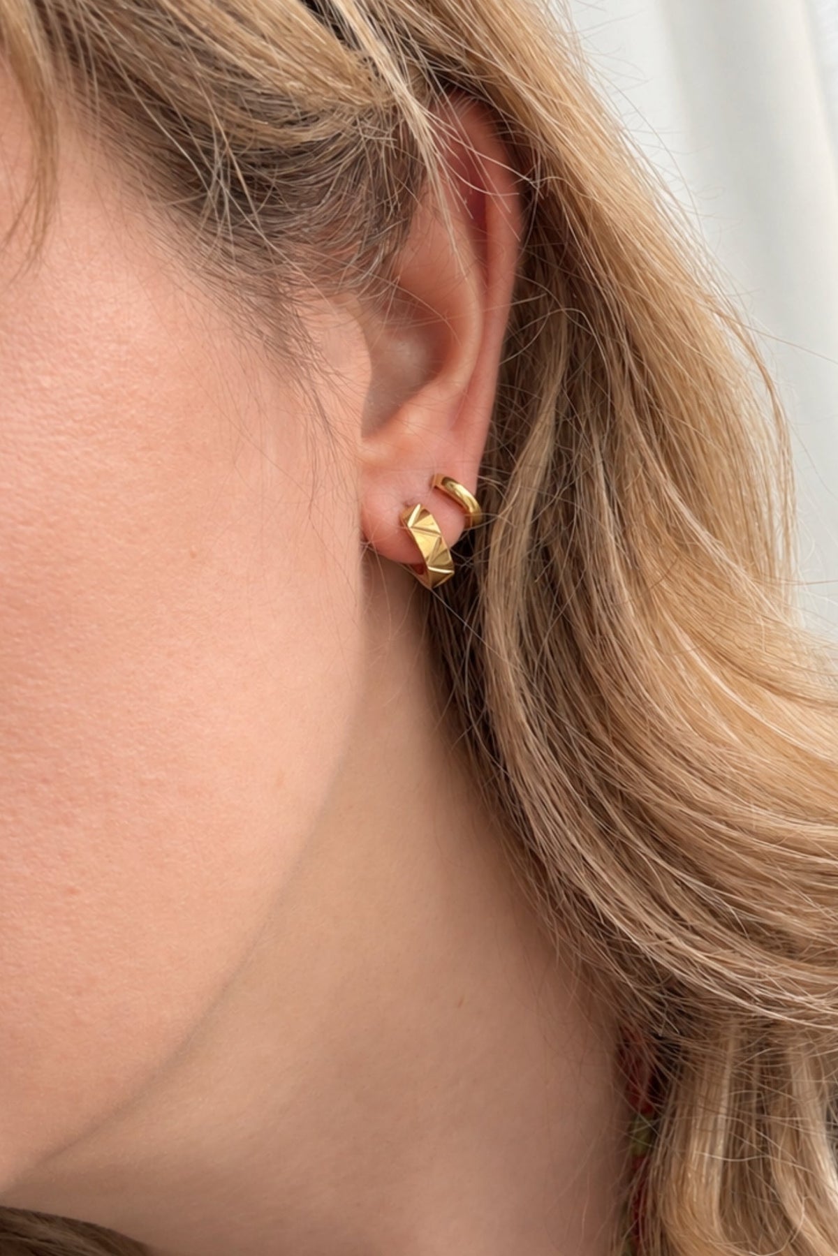 A close-up side profile of a woman with blonde hair wearing a small gold huggie hoop earring.
