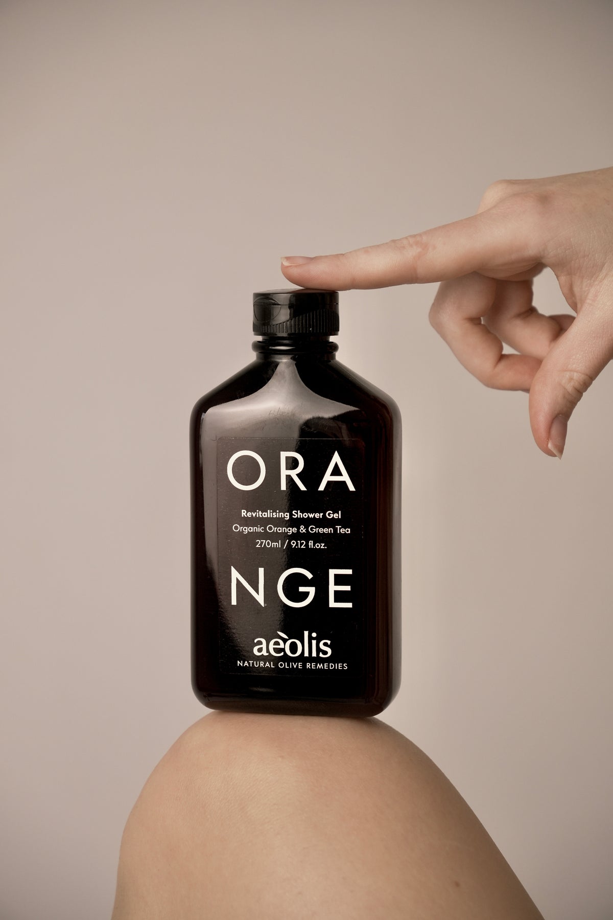 A bottle of aeolis Orange Nge Revitalising Shower Gel sits on a person's knee, with a hand pointing at the label. The bottle is dark brown glass with white text, set against a solid beige background.