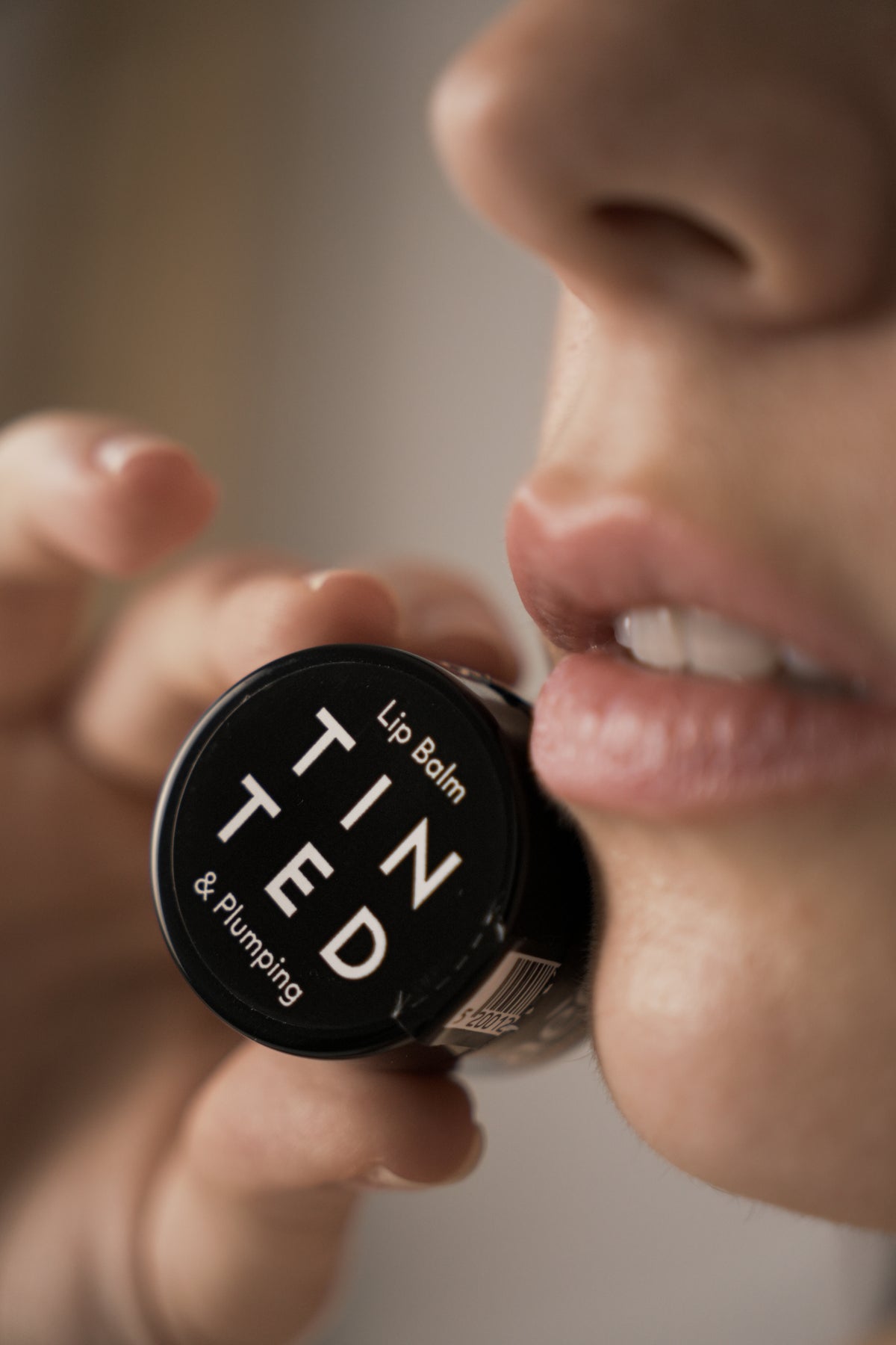 A close-up lifestyle shot of a person holding a black lip balm container to their lips. The container has a black lid with white text that reads 'TIN TED Lip Balm & Plumping'.