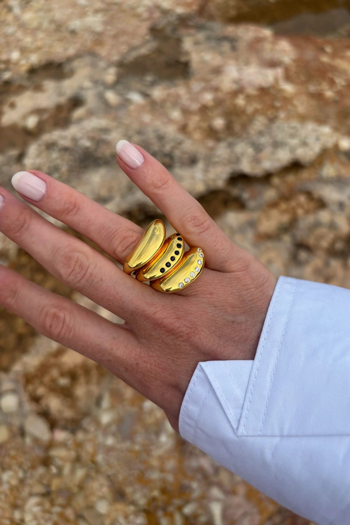 A close-up of a hand wearing three stacked gold rings with textured, organic shapes, set against a blurred rocky outdoor background.