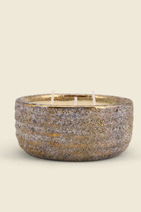 A three-wick candle in a textured, ribbed ceramic bowl with a speckled gold, silver, and gray exterior and a gold-toned interior, set against a solid cream background.