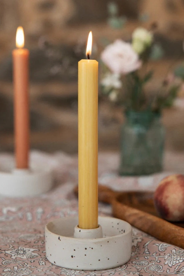 Yellow candle in a white holder on a patterned tablecloth with peaches and flowers in the background.