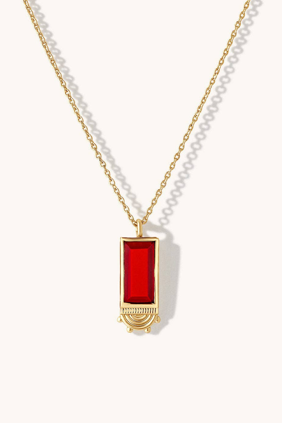 A gold chain necklace featuring a rectangular red gemstone pendant with a gold crescent moon design at the bottom, displayed on a clean off-white background.