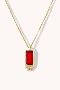 A gold chain necklace featuring a rectangular red gemstone pendant with a gold crescent moon design at the bottom, displayed on a clean off-white background.