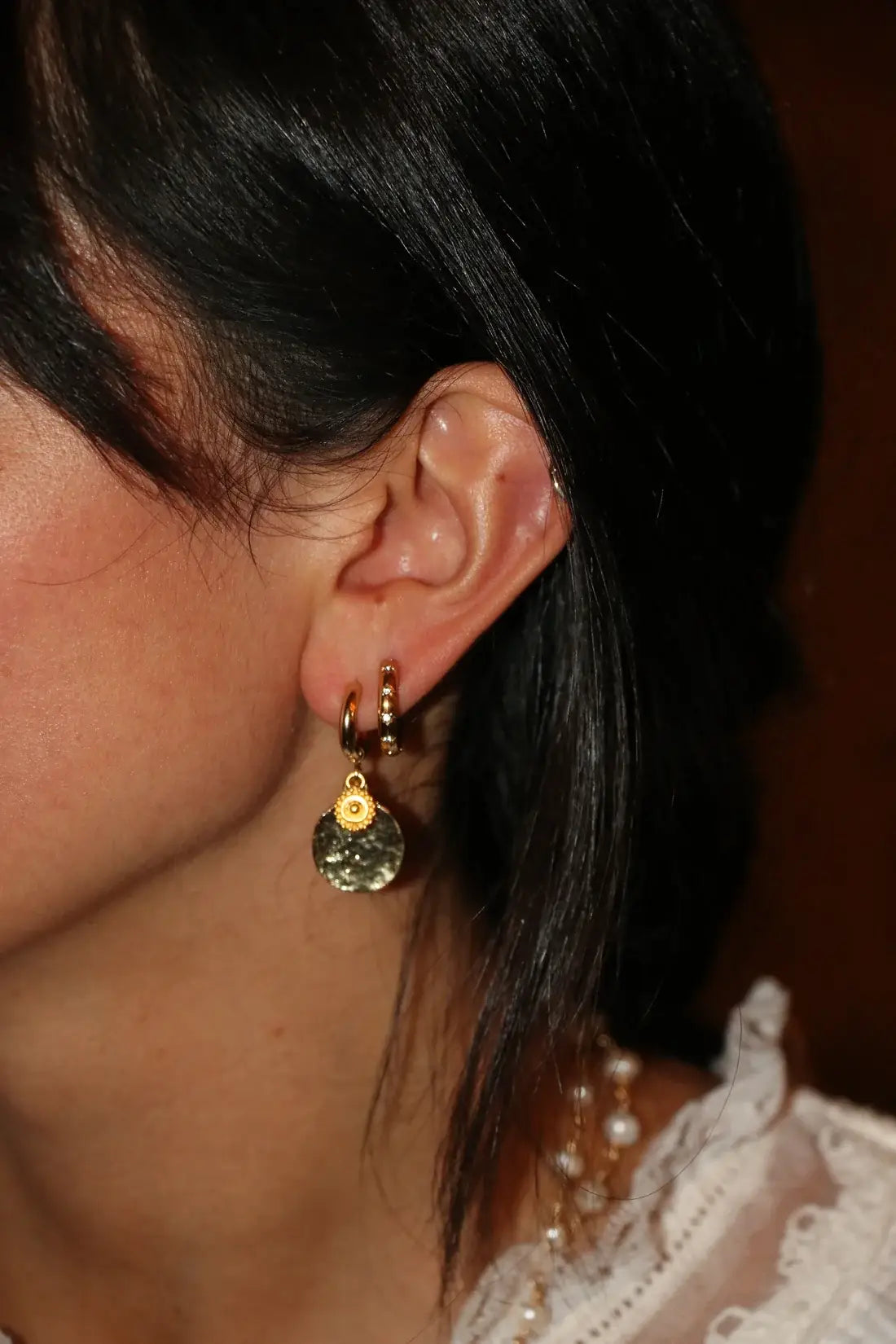 A close-up of a person's ear wearing a gold huggie hoop earring with a small gold disc charm and a dangling textured round charm in a dark greenish tone.