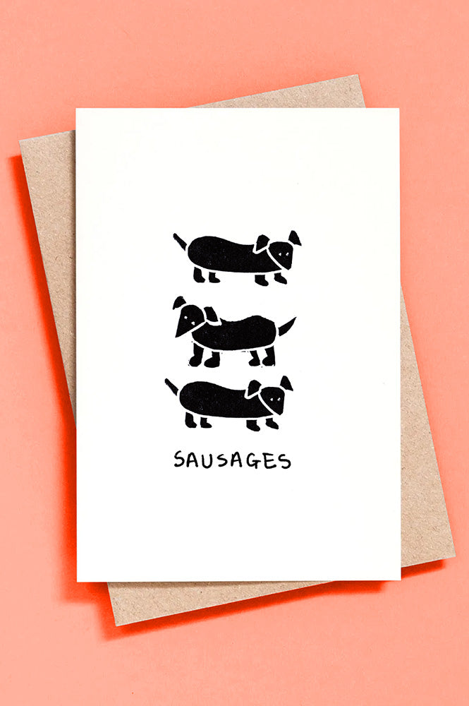 Greeting card with black illustrations of sausage dogs on a white background, placed on a pink surface.