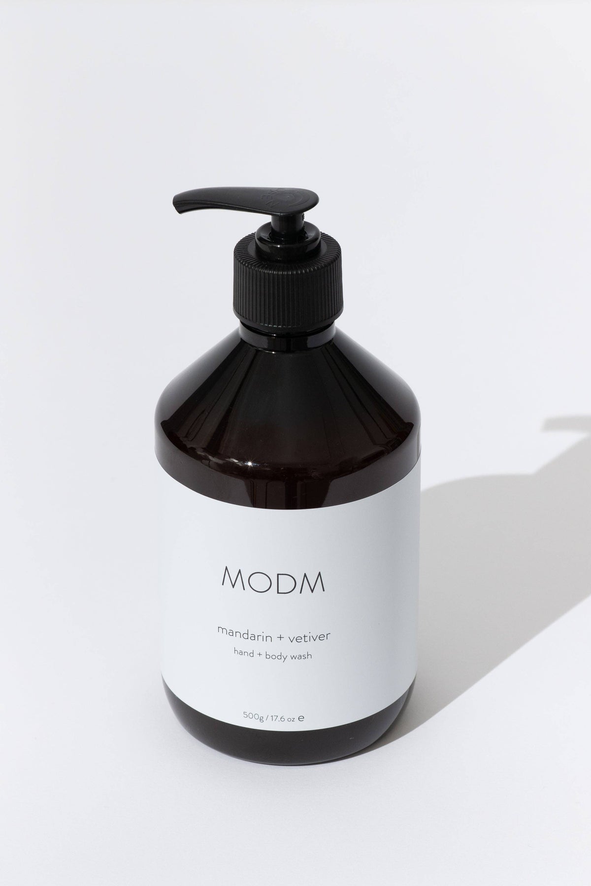 A dark amber glass pump bottle of MODM hand and body wash with a white label, set against a clean white background with a soft shadow.