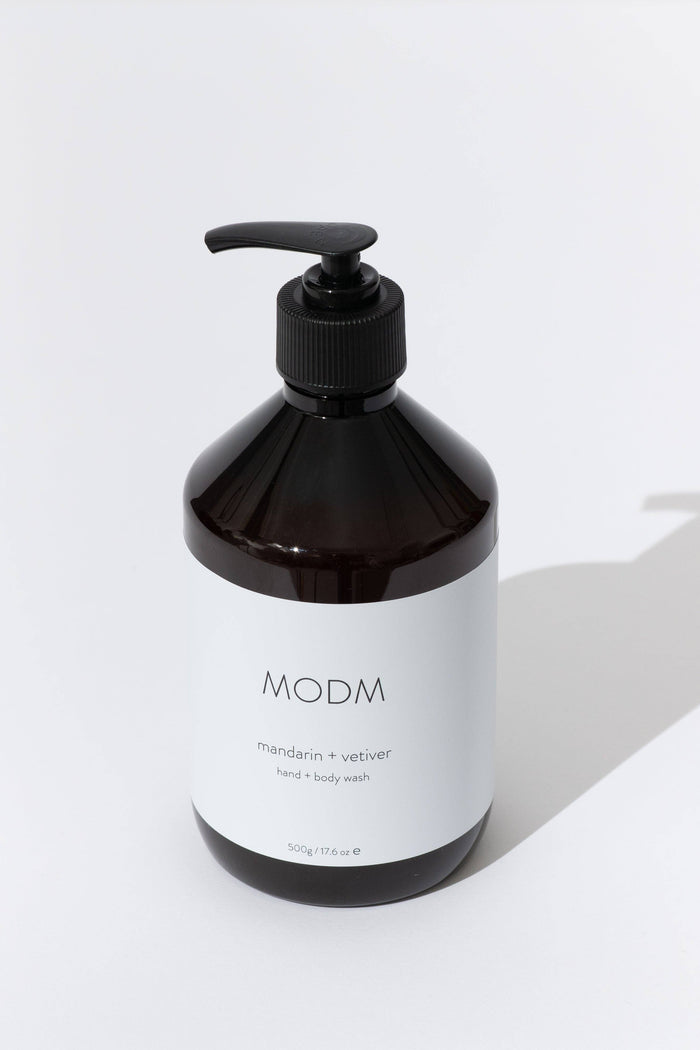 A dark amber glass pump bottle of MODM hand and body wash with a white label, set against a clean white background with a soft shadow.