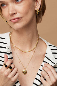 Woman wearing gold jewelry including a necklace, earrings, and rings on a beige background
