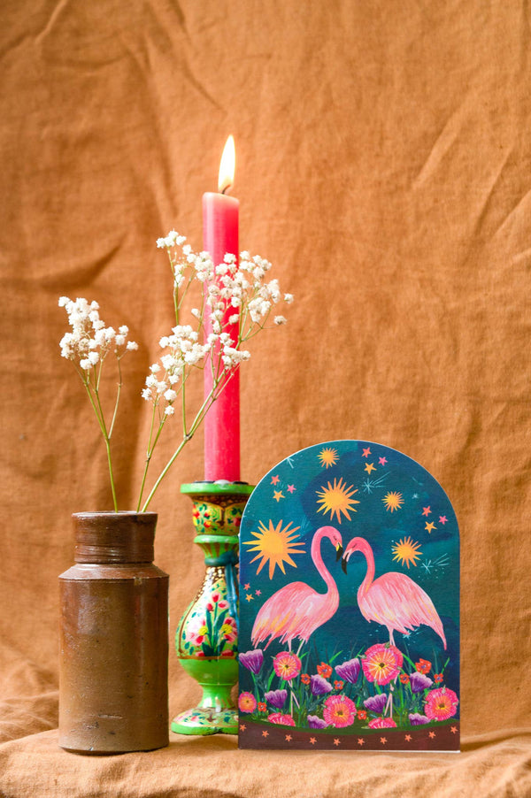 Decorative card with flamingo design next to a lit candle and vase on a brown fabric background