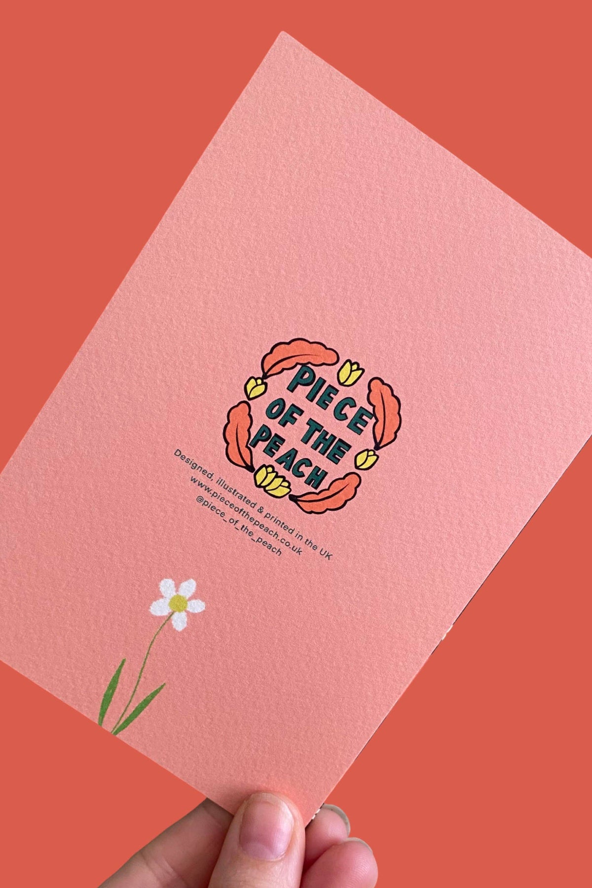 A hand holds a textured pink card featuring a retro-style illustration of a flower and the text 'PIECE OF THE BEACH' against a solid coral background.