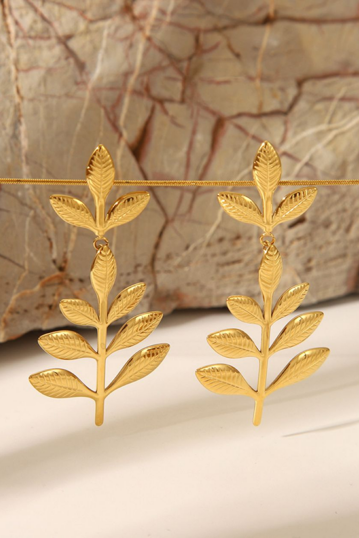 A pair of gold leaf dangle earrings hanging from a thin gold wire, set against a textured beige and brown stone background.