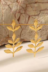 A pair of gold leaf dangle earrings hanging from a thin gold wire, set against a textured beige and brown stone background.