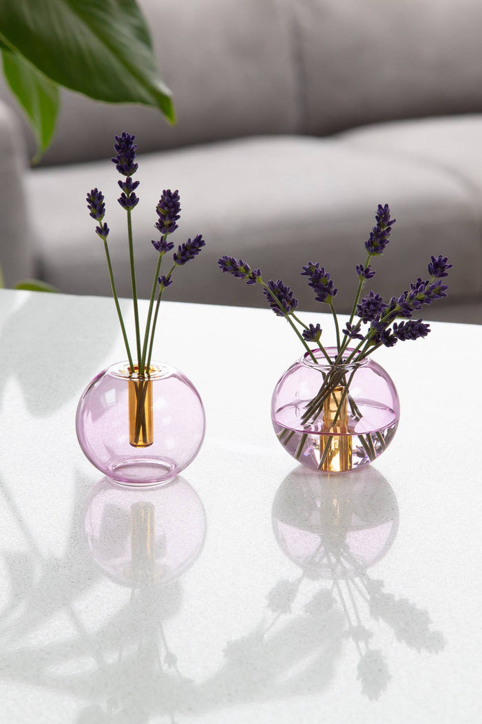 Two small round glass vases with lavender on a reflective surface.