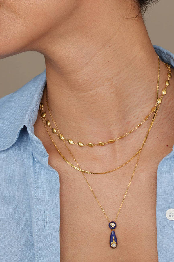A close-up of a model wearing three layered gold necklaces, including a teardrop pendant with a dark blue and white stone, over a light blue collared shirt.