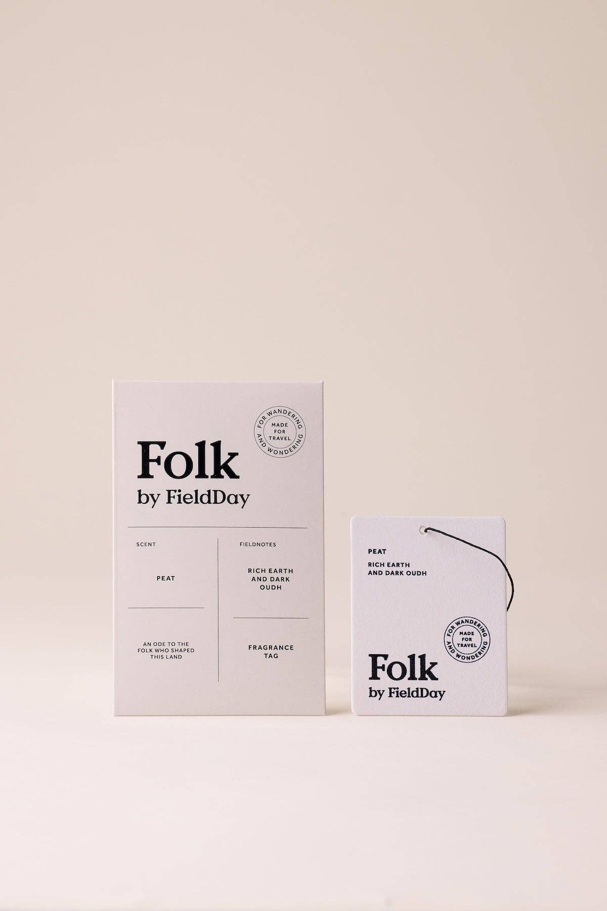A minimalist fragrance product setup on a warm off-white background, featuring a tall rectangular box and a smaller hanging tag both printed with the brand name 'Folk by FieldDay' in black text.
