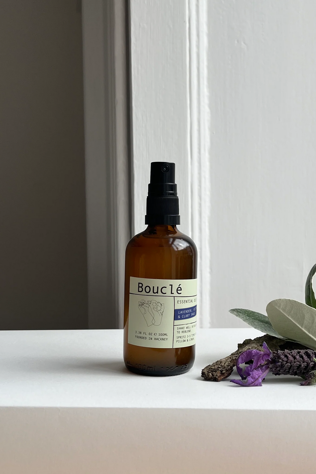 An amber glass spray bottle labeled Bouclé Essential stands on a white windowsill with dried lavender, green leaves, and purple flowers beside it. The background shows a white paneled window frame and a dark gray wall.