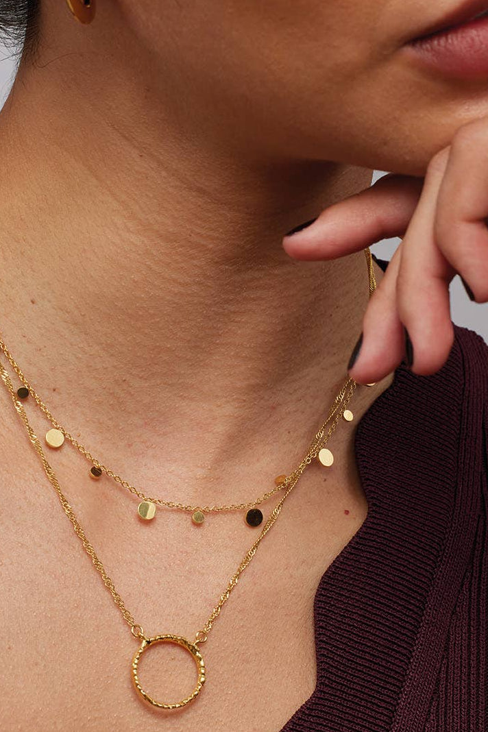 A close-up of a model wearing a layered gold necklace set, featuring delicate chains with small gold and black round charms and a textured gold ring pendant, styled with a burgundy ribbed top.