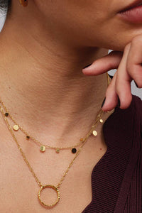 A close-up of a model wearing a layered gold necklace set, featuring delicate chains with small gold and black round charms and a textured gold ring pendant, styled with a burgundy ribbed top.